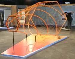 A modern art sculpture made of orange metal rods forming geometric and curved shapes on a raised platform, displayed in an indoor exhibition space.