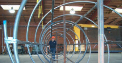 A person wearing a hard hat works on a large, circular metal framework inside a warehouse with exposed beams and industrial equipment.